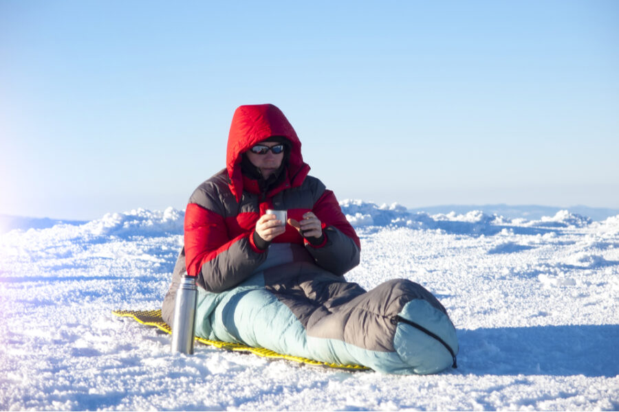 man sitting on a sleeping bag drinking hot coffee from a thermos